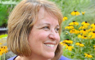 Woman with short light brown hair smiling. Yellow flowers and greenery sit behind her.